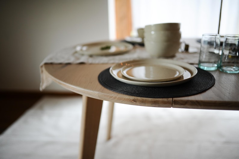 May include: A round light-colored wooden dining table featuring a black circular placemat, a set of stacked cream-colored ceramic plates, and two clear glass tumblers. A light-colored table runner and additional stacked bowls are visible.