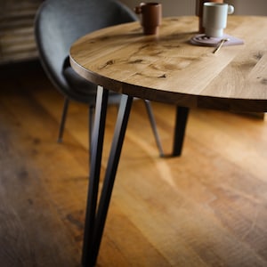 May include: A round wooden dining table with black metal legs. The tabletop is made of light-colored wood and has a natural grain pattern. Two brown ceramic mugs and a white mug sit on the table. A gray chair is in the background.