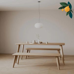 Könnte beinhalten: Ein heller Holzesstisch mit zwei passenden Bänken. Auf dem Tisch stehen eine weiße Teekanne, eine kleine Vase mit einem Zweig und zwei kleine Tassen. Eine weiße Pendelleuchte hängt über dem Tisch und eine orangefarbene Ornamentgrafik ist oben rechts.