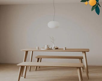 Op de afbeelding: Een lichtgekleurde houten eettafel met twee bijpassende banken. Op de tafel staan een witte theepot, een klein vaasje met een takje en twee kleine kopjes. Een witte hanglamp hangt boven de tafel en een oranje ornamentafbeelding staat rechtsboven.