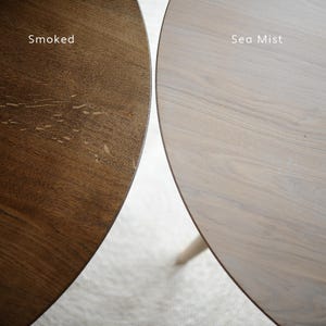 Puede incluir: Primer plano de dos tableros de mesa redondos de madera. Uno es marr&oacute;n oscuro, etiquetado como "Smoked", y el otro es gris claro, etiquetado como "Sea Mist". La veta de la madera es visible en ambas superficies, mostrando la textura natural y las variaciones de color.
