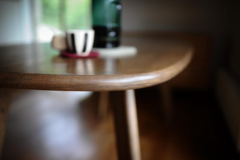 May include: Close-up of a wooden table with a curved edge. A white cup with black stripes rests on a red coaster. A green bottle is in the background. The table has a natural wood grain.