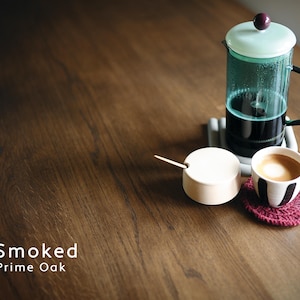 May include: A French press coffee maker with a light green lid and a black handle sits on a gray coaster. A small white sugar bowl with a wooden spoon and a cup of coffee with a black and white design are on a wooden table. A red coaster is under the coffee cup. The text "Smoked Prime Oak" is at the bottom.