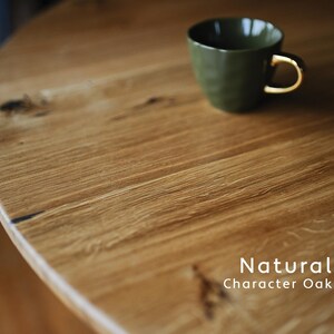 Puede incluir: Una mesa redonda de roble natural con vetas y nudos visibles, con una taza de cer&aacute;mica verde oliva oscuro con asa dorada encima. El texto Natural Character Oak est&aacute; impreso en blanco en la esquina inferior derecha de la imagen.