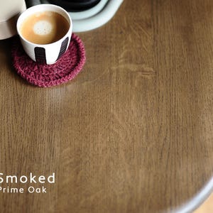 May include: A curved wooden tabletop with a Smoked Prime Oak finish, featuring a visible wood grain texture. A small white cup with black vertical stripes containing coffee sits on a round, dark red crocheted coaster on the surface.