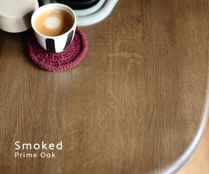 May include: A curved wooden tabletop with a Smoked Prime Oak finish, featuring a visible wood grain texture. A small white cup with black vertical stripes containing coffee sits on a round, dark red crocheted coaster on the surface.