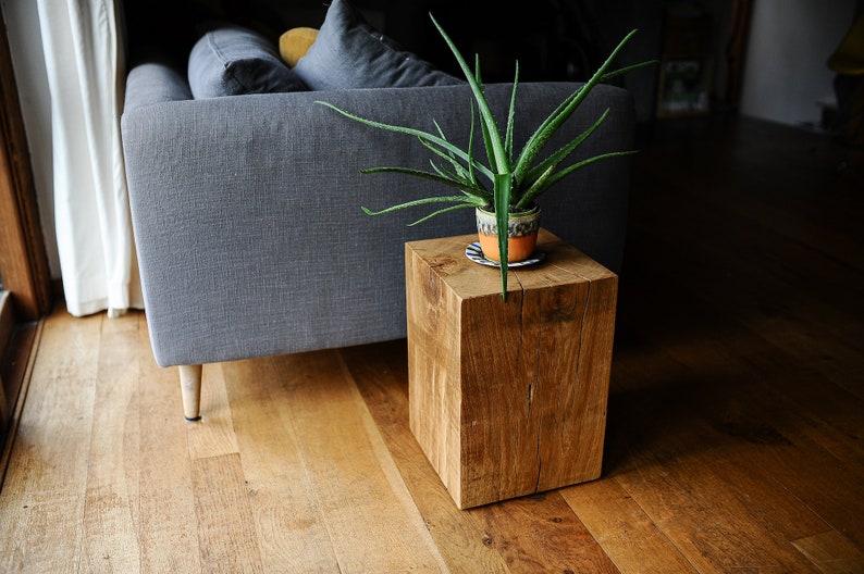 May include: A wooden end table with a natural finish and a plant in a terracotta pot on top. The table is in front of a gray sofa.
