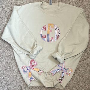 May include: A beige sweatshirt with a large patchwork monogram on the front and two smaller patchwork bows on the sleeves.