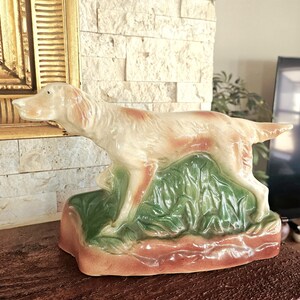 May include: A ceramic figurine of a white and brown dog standing on a green and brown base. The dog is in a hunting pose, with its head turned to the side and its tail raised.