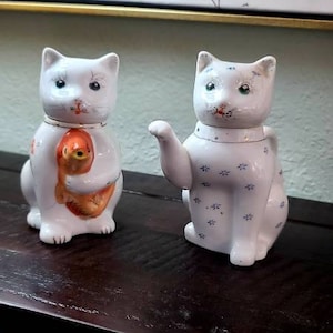 May include: Two white porcelain cat figurines. One cat is holding a small orange fish and the other cat is a teapot with a blue and white floral pattern.