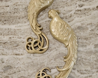 May include: Two gold-colored, decorative bird wall sculptures. Each bird has detailed feather and tail designs, with a curved, ornate base. The birds are positioned on a light-colored, textured surface.