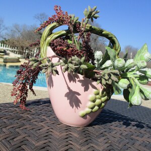 May include: A pink ceramic planter with a unique, wavy design. The planter is filled with a variety of green and brown plants, including succulents and eucalyptus.