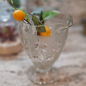 May include: A clear glass pitcher with a textured surface and floral design. A small orange citrus fruit is inside the pitcher.