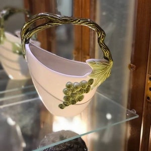 May include: A white ceramic bowl with a green vine handle and green grapes. The bowl is displayed on a glass shelf in a wooden cabinet.