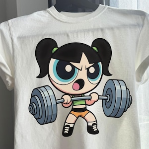 Könnte beinhalten: Weißes T-Shirt mit einer Zeichentrickfigur, die eine Langhantel hebt. Die Figur hat schwarze Haare, blaue Augen und trägt ein grünes Oberteil und orangefarbene Shorts. Die Langhantel ist grau. Das T-Shirt hängt an einem schwarzen Kleiderbügel.