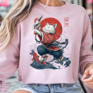 Może przedstawiać: Jasnoróżowa bluza z nadrukiem białego kota w tradycyjnym japońskim stroju, trzymającego łuk. Projekt zawiera czerwone słońce, niebieskie fale i japoński tekst. Tekst "LIGHT PINK" znajduje się w jasnoróżowym polu w lewym górnym rogu.