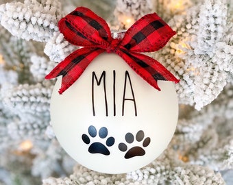 Puede incluir: Una bola de Navidad blanca con el nombre "MIA" en letras negras y dos huellas de patas. Está rematada con un lazo a cuadros rojo y negro. La bola cuelga de un árbol de Navidad cubierto de nieve.
