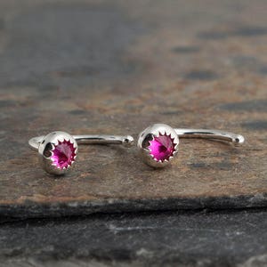 May include: A pair of silver nose rings with a pink gemstone in the center of each ring.