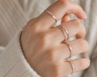 May include: A hand wearing several thin, silver rings. The rings have a textured, twisted design. The hand is wearing a white sweater. The rings are a simple, minimalist style.