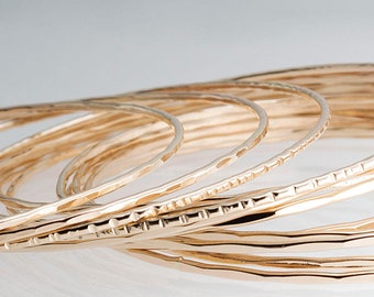 Könnte beinhalten: Eine Sammlung goldfarbener Armreifen mit strukturierten Oberflächen. Die Armreifen haben ein gehämmertes oder geätztes Design, das einen einzigartigen und eleganten Look erzeugt. Der Schmuck wird vor einem weichen, neutralen Hintergrund präsentiert.