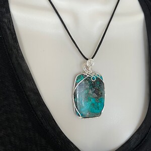 Puede incluir: Un colgante de piedra turquesa y negra con envoltura de alambre plateado, colgado de un collar de cord&oacute;n negro.