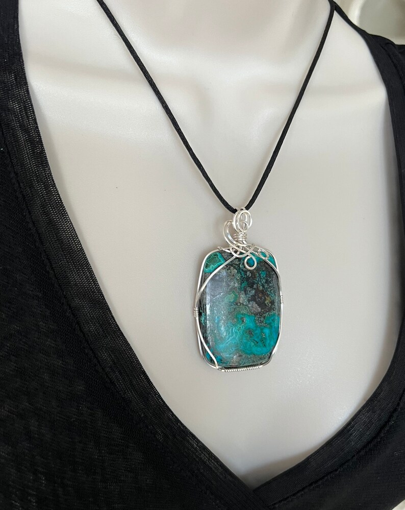 Puede incluir: Un colgante de piedra turquesa y negra con envoltura de alambre plateado, colgado de un collar de cord&oacute;n negro.