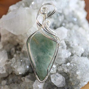 May include: A silver wire wrapped pendant featuring a green gemstone. The pendant is displayed on a bed of white crystals.