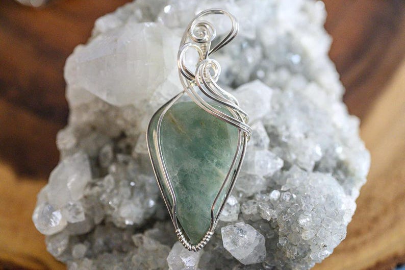 May include: A silver wire wrapped pendant featuring a green gemstone. The pendant is displayed on a bed of white crystals.