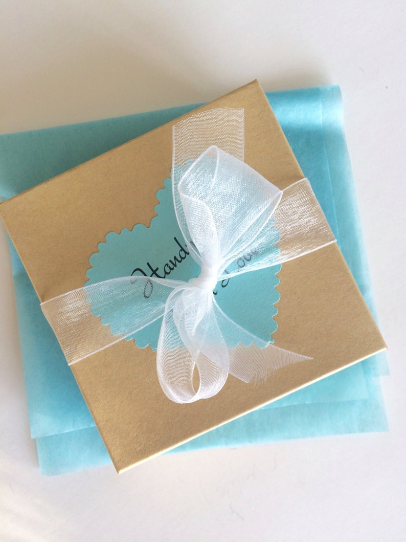 May include: A gold gift box with a white ribbon tied in a bow. The box is sitting on a light blue tissue paper. The box has a blue heart-shaped sticker with the text "Handmade!" on it.