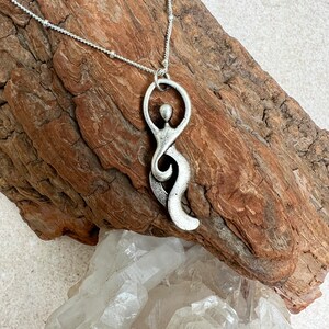 Pu&ograve; includere: Una collana d'argento con un ciondolo a forma di figura umana stilizzata. Il ciondolo &egrave; astratto e presenta un design fluido e organico. La collana &egrave; su una superficie di legno con un gruppo di cristalli di quarzo trasparenti.