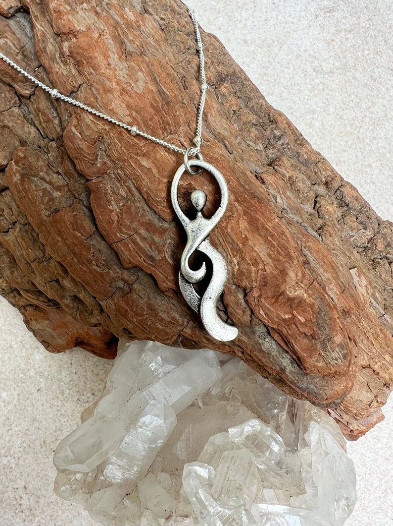 Pu&ograve; includere: Una collana d'argento con un ciondolo a forma di figura umana stilizzata. Il ciondolo &egrave; astratto e presenta un design fluido e organico. La collana &egrave; su una superficie di legno con un gruppo di cristalli di quarzo trasparenti.