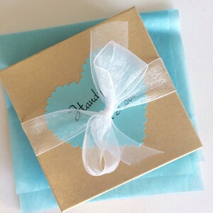 May include: A gold gift box with a white ribbon tied in a bow. The box is sitting on a light blue tissue paper. The box has a blue heart-shaped sticker with the text "Handmade!" on it.