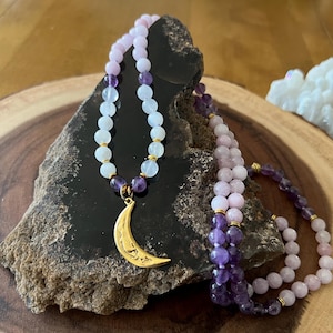 Könnte beinhalten: Eine Halskette mit einem goldenen Halbmond-Anhänger, mit hellvioletten, weißen und Amethyst-Perlen. Die Halskette liegt auf einem dunklen Stein, mit einem weißen Kristallcluster im Hintergrund.