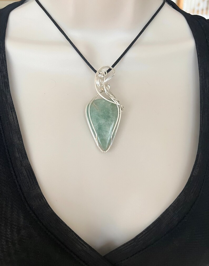 May include: A silver wire wrapped pendant featuring a green gemstone. The pendant is hanging from a black cord.