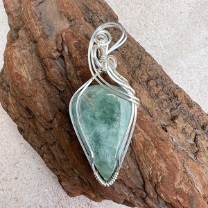 May include: A silver wire wrapped pendant featuring a green and white fluorite gemstone. The pendant is displayed on a piece of driftwood.
