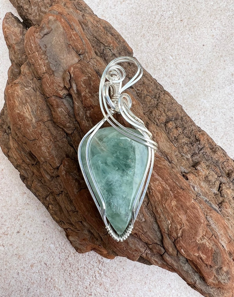 May include: A silver wire wrapped pendant featuring a green and white fluorite gemstone. The pendant is displayed on a piece of driftwood.