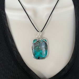 Puede incluir: Un colgante de piedra turquesa y verde con envoltura de alambre plateado, colgado de un collar de cord&oacute;n negro.