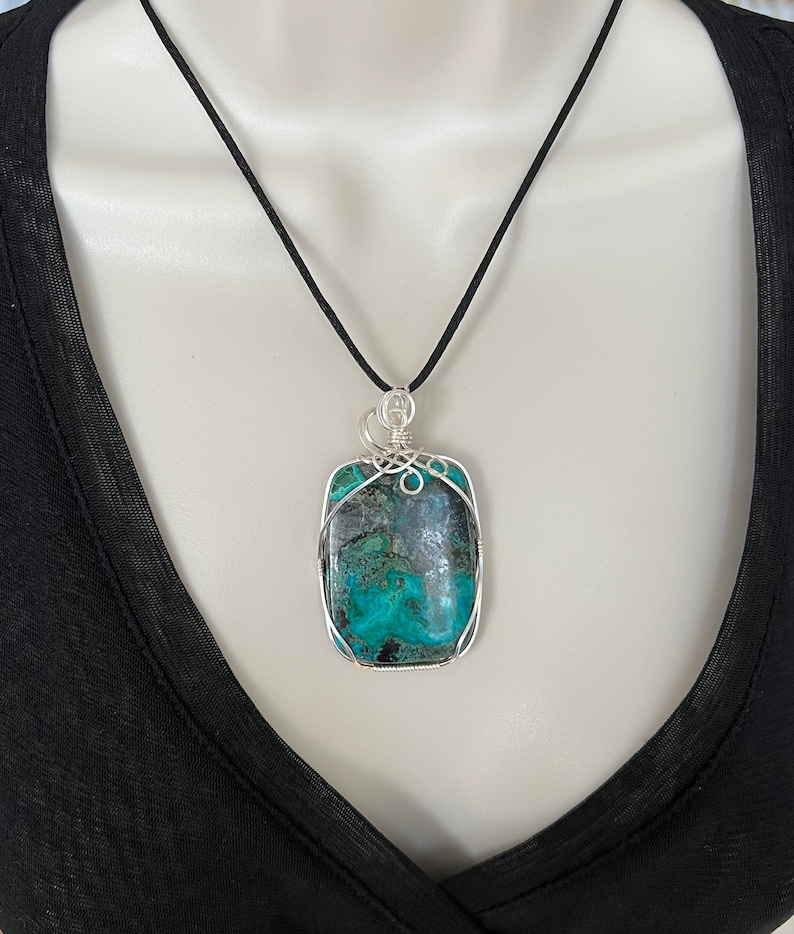 Puede incluir: Un colgante de piedra turquesa y verde con envoltura de alambre plateado, colgado de un collar de cord&oacute;n negro.