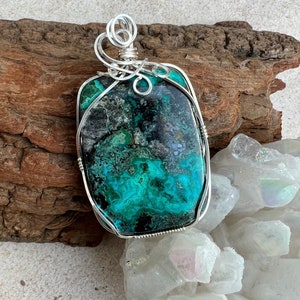 Puede incluir: Un colgante de alambre plateado envuelto alrededor de una gran piedra preciosa rectangular verde y negra. La piedra preciosa tiene un patr&oacute;n de remolino y m&aacute;rmol. El colgante se muestra en un trozo de madera a la deriva y una cama de cristales blancos.