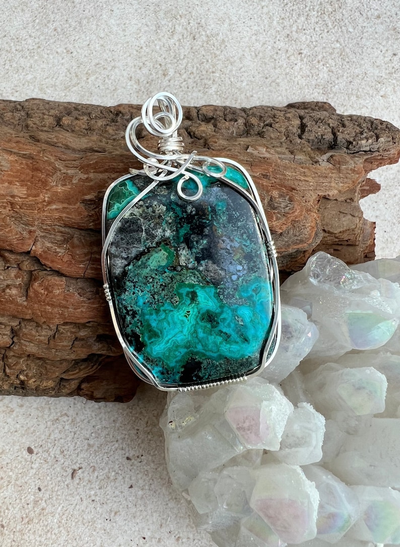 Puede incluir: Un colgante de alambre plateado envuelto alrededor de una gran piedra preciosa rectangular verde y negra. La piedra preciosa tiene un patr&oacute;n de remolino y m&aacute;rmol. El colgante se muestra en un trozo de madera a la deriva y una cama de cristales blancos.