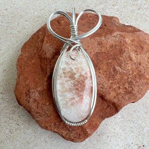 May include: A silver wire wrapped pendant featuring a light pink and white oval shaped stone with a swirling pattern. The pendant is hanging from a silver wire heart shaped bail.