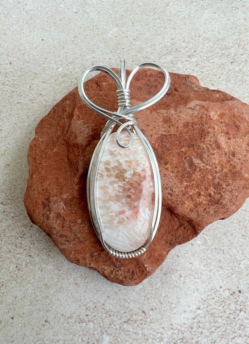 May include: A silver wire wrapped pendant featuring a light pink and white oval shaped stone with a swirling pattern. The pendant is hanging from a silver wire heart shaped bail.
