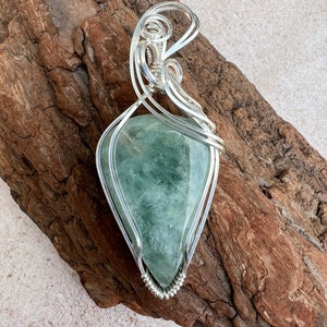 May include: A green gemstone pendant wrapped in silver wire. The pendant is shaped like a teardrop and has a delicate, intricate design.