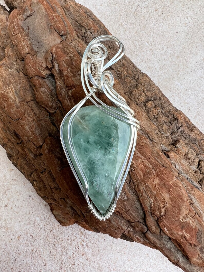 May include: A green gemstone pendant wrapped in silver wire. The pendant is shaped like a teardrop and has a delicate, intricate design.