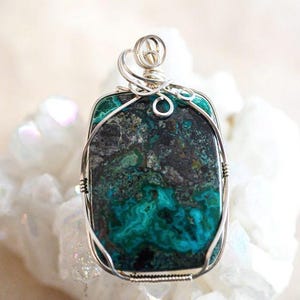 Puede incluir: Un colgante de alambre plateado envuelto alrededor de una piedra de crisocola verde y negra. La piedra tiene un patr&oacute;n veteado &uacute;nico.