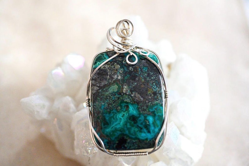 Puede incluir: Un colgante de alambre plateado envuelto alrededor de una piedra de crisocola verde y negra. La piedra tiene un patr&oacute;n veteado &uacute;nico.