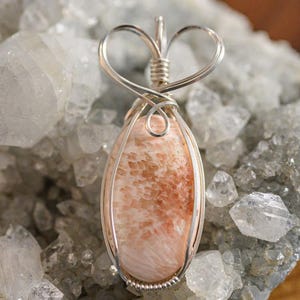 May include: A pink and orange gemstone pendant with a heart-shaped wire wrap. The pendant is set against a background of white crystals.