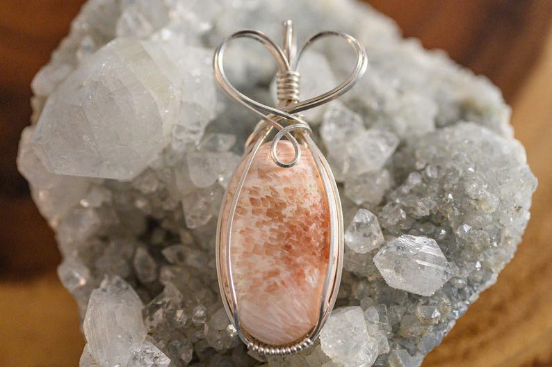 May include: A pink and orange gemstone pendant with a heart-shaped wire wrap. The pendant is set against a background of white crystals.