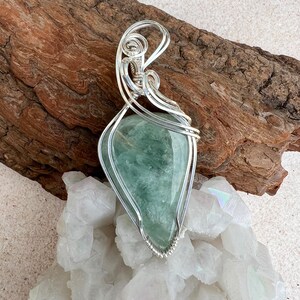 May include: A silver wire wrapped pendant featuring a light green gemstone. The pendant is displayed on a bed of white crystals and a piece of driftwood.