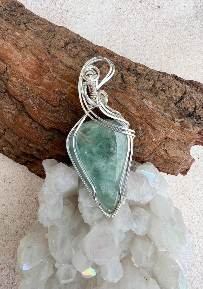 May include: A silver wire wrapped pendant featuring a light green gemstone. The pendant is displayed on a bed of white crystals and a piece of driftwood.